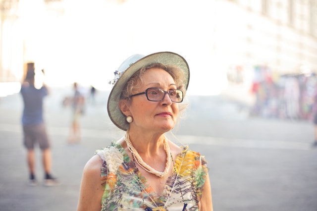 Staršia žena vo farebných šatách a klobúku s výrazným náhrdelníkom a náušnicami.jpg